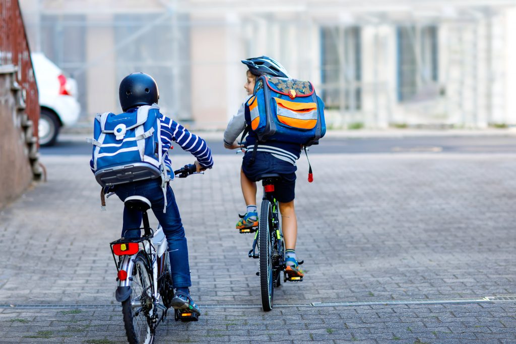 Two School Kid Boys In Safety Helmet Riding With Bike In The City With Backpacks. Happy Children In Colorful Clothes Biking On Bicycles On Way To School. Safe Way For Kids Outdoors To School